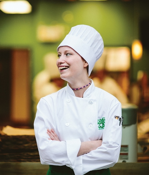 Young woman in a white chef's jacket and chefs hat, looking to the left and smiling